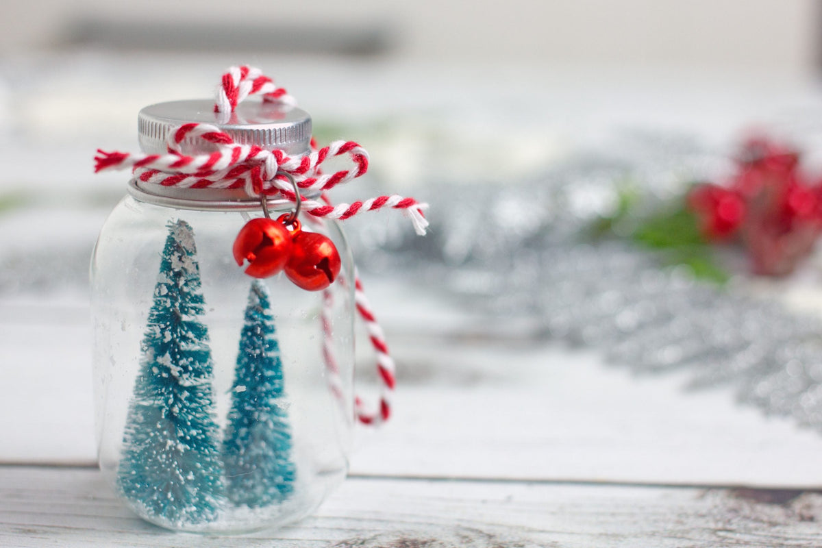 A decorated gift jar with small evergreen trees, red bells, and red-and-white ribbon.