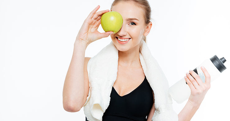 A woman holds a green apple near her eye and a water bottle in her other hand.
