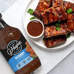 A bottle of G. Hughes Zero Sugar Hickory BBQ Sauce is displayed next to a plate of glazed ribs garnished with parsley and a small bowl of BBQ sauce on a light background.