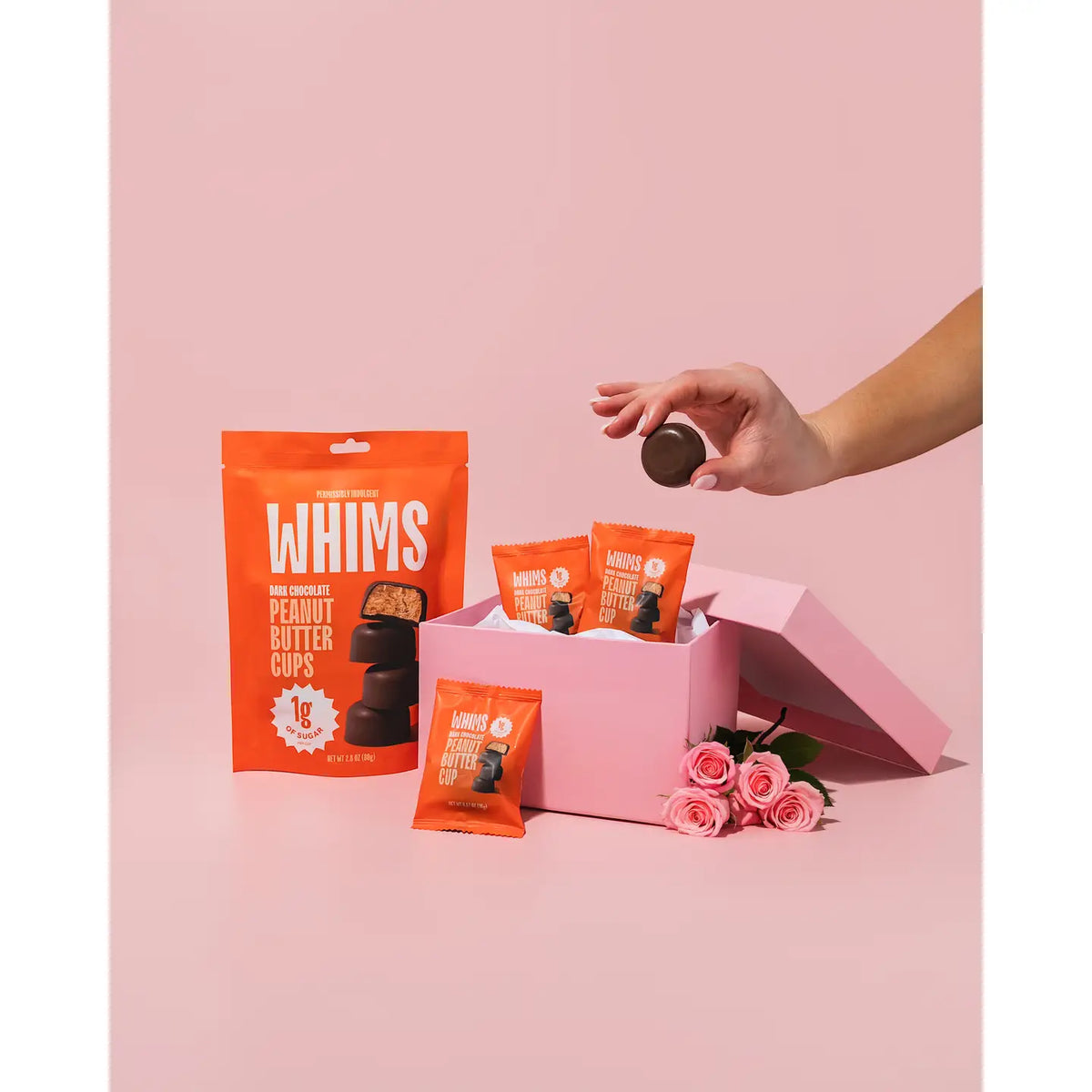 A hand is reaching for a dark chocolate peanut butter cup from a pink gift box filled with more cups and surrounded by roses, set against a soft pink background.