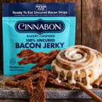 A package of Cinnabon-inspired 100% uncured bacon jerky is displayed alongside a frosted cinnamon roll and cinnamon sticks on a rustic wooden surface.