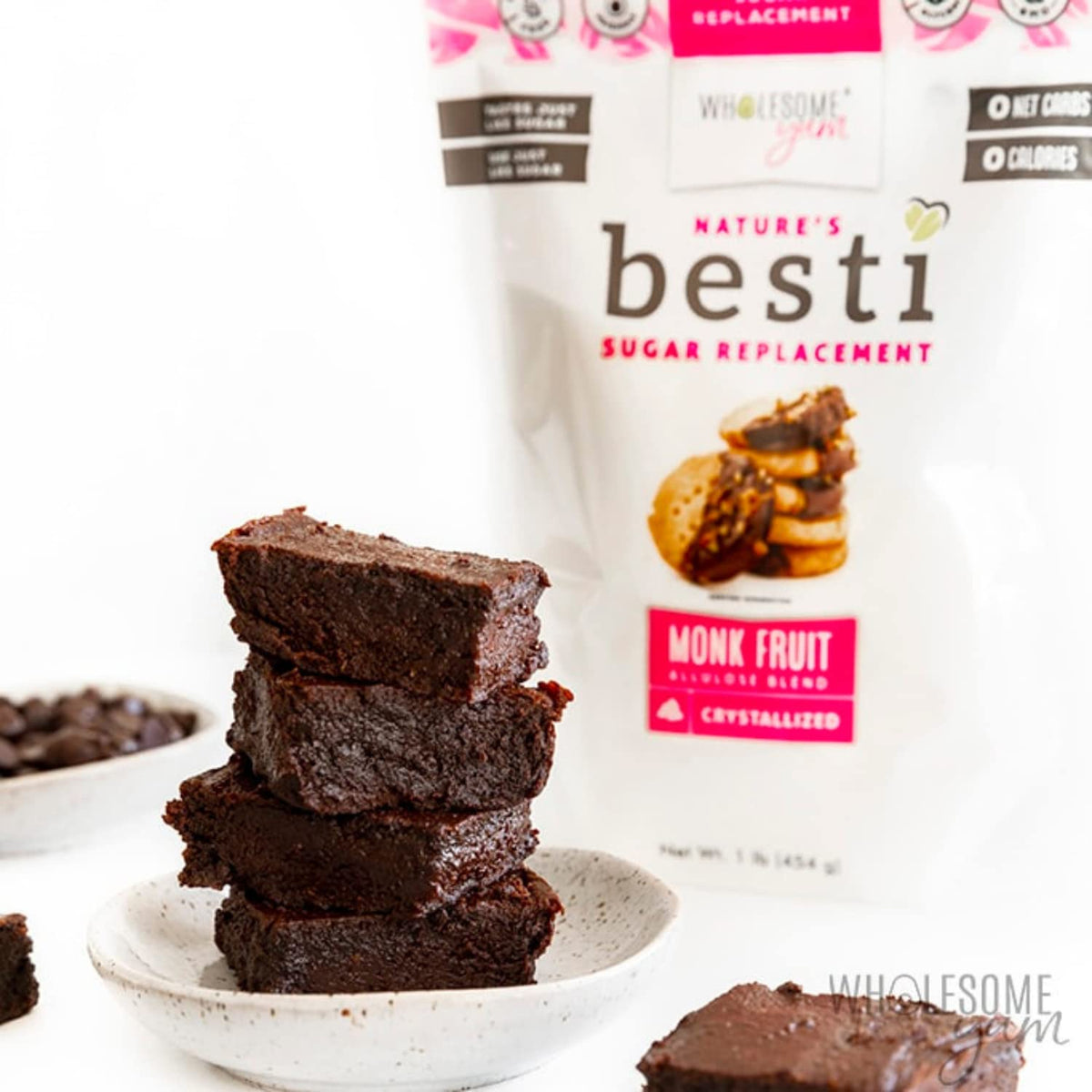 A stack of rich, dark brownies sits on a small plate, with a bag of Wholesome Yum's Besti Monk Fruit Sugar Replacement in the background, surrounded by chocolate chips on a white surface.