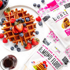 A golden-brown waffle topped with fresh strawberries, blueberries, and raspberries sits on a plate beside packages of almond flour and sugar replacement, with a small glass of syrup nearby.