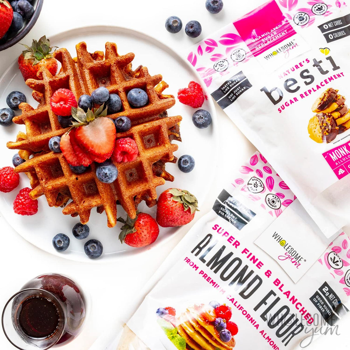 A golden-brown waffle topped with fresh strawberries, blueberries, and raspberries sits on a plate beside packages of almond flour and sugar replacement, with a small glass of syrup nearby.