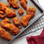 Crispy, golden-brown fried chicken tenders are arranged on a baking tray, showcasing their crunchy coating, with a red kitchen towel partially visible in the corner.