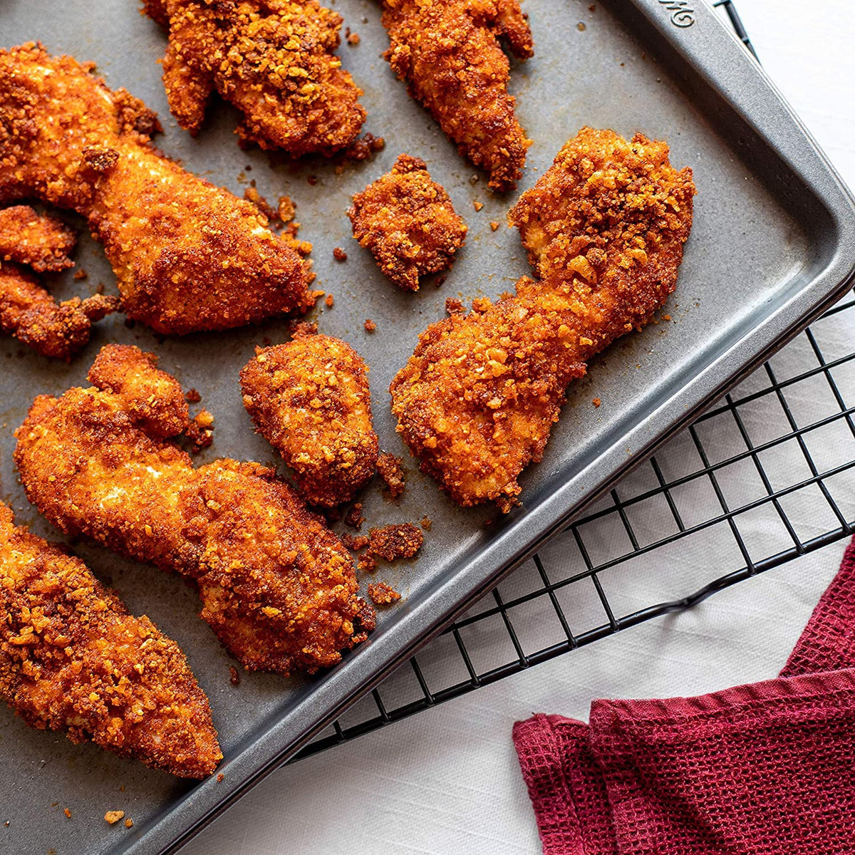 Crispy, golden-brown fried chicken tenders are arranged on a baking tray, showcasing their crunchy coating, with a red kitchen towel partially visible in the corner.