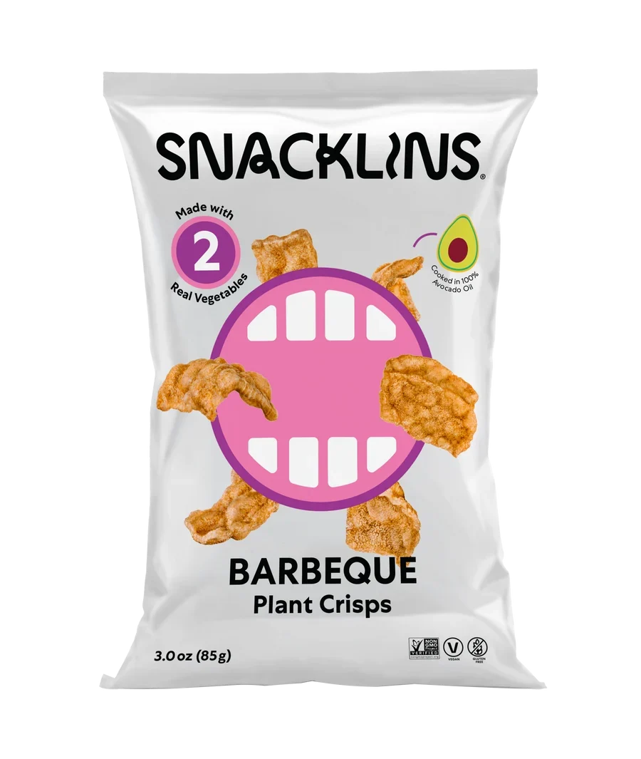 A bag of Snacklins Barbeque Plant Crisps features a playful design with a large pink mouth graphic, showcasing crispy snacks made from real vegetables and highlighting their avocado oil content.