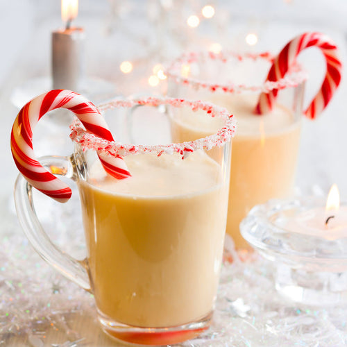 Two clear mugs filled with creamy holiday drinks are garnished with crushed peppermint and topped with red and white striped candy canes, set against a softly lit, festive background.