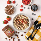 A bowl of oatmeal topped with sliced strawberries and drizzled chocolate hazelnut spread is surrounded by fresh strawberries, raspberries, blueberries, a jar of hazelnut spread, and black spoons on a checkered cloth.