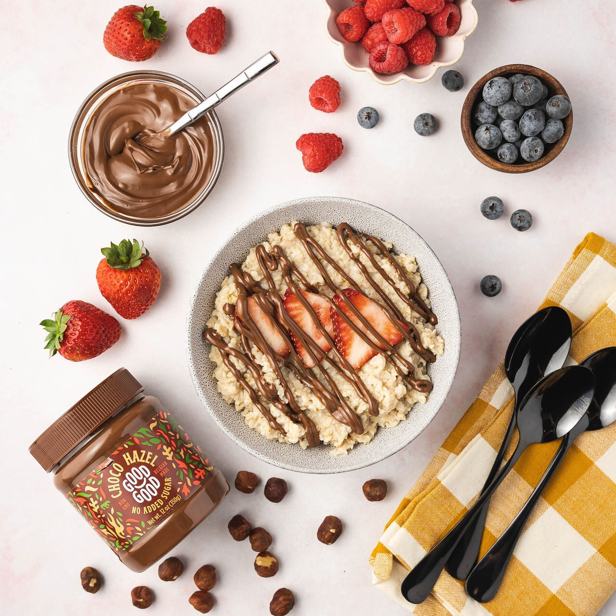 A bowl of oatmeal topped with sliced strawberries and drizzled chocolate hazelnut spread is surrounded by fresh strawberries, raspberries, blueberries, a jar of hazelnut spread, and black spoons on a checkered cloth.