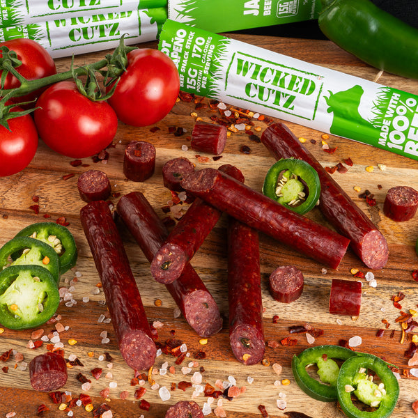 A wooden cutting board displays various pieces of beef jerky, sliced jalapeños, whole tomatoes, and chili flakes, with packaging for "Wicked Cutz" visible in the background.
