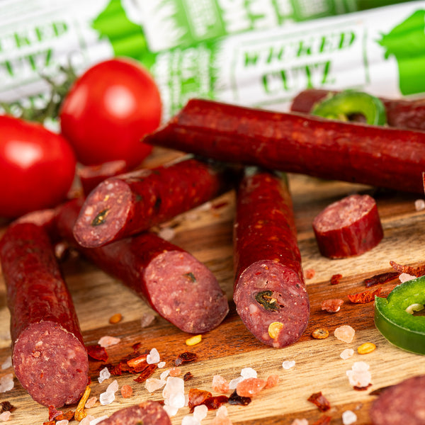 A wooden surface displays several pieces of spicy beef jerky, some cut in half to reveal their texture, surrounded by fresh tomatoes, jalapeño slices, and scattered spices.