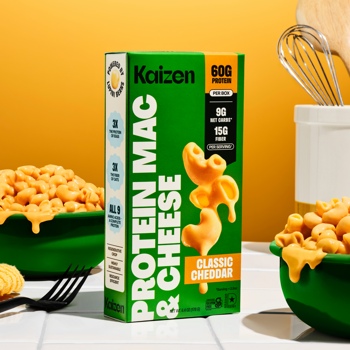 A colorful box of Kaizen Protein Mac & Cheese sits next to a green bowl filled with cheesy pasta, against a bright orange background, highlighting its nutritional benefits and appealing presentation.