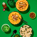 Three bowls of pasta dishes are arranged on a bright green surface, featuring creamy macaroni with chicken, a spinach and cheese blend, and condiments like chili flakes and a green drink with ice.