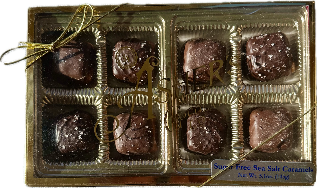 A gold box tied with a ribbon contains eight chocolate-covered caramels sprinkled with sea salt, labeled as "Sugar Free Sea Salt Caramels" from Asher's Chocolate Co.