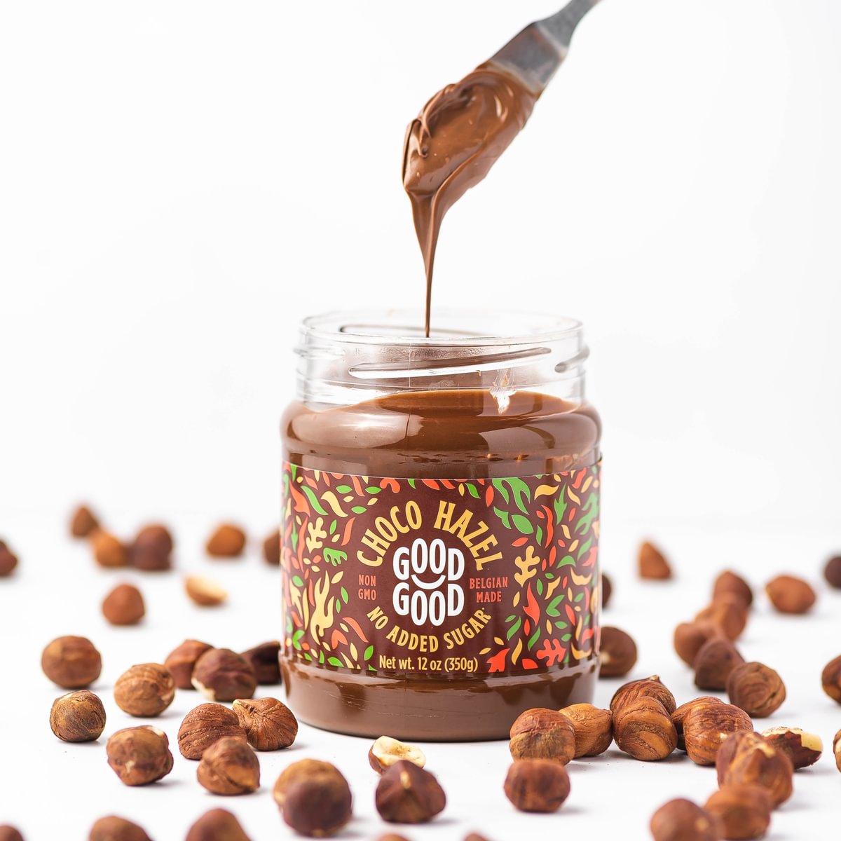 A jar of chocolate hazelnut spread with a spoon dripping the creamy texture is surrounded by scattered hazelnuts on a white background.