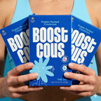 A person holds three boxes of "Boost Couscous," featuring bright blue packaging with playful graphics, showcasing the product's protein and gluten-free attributes against a light blue background.