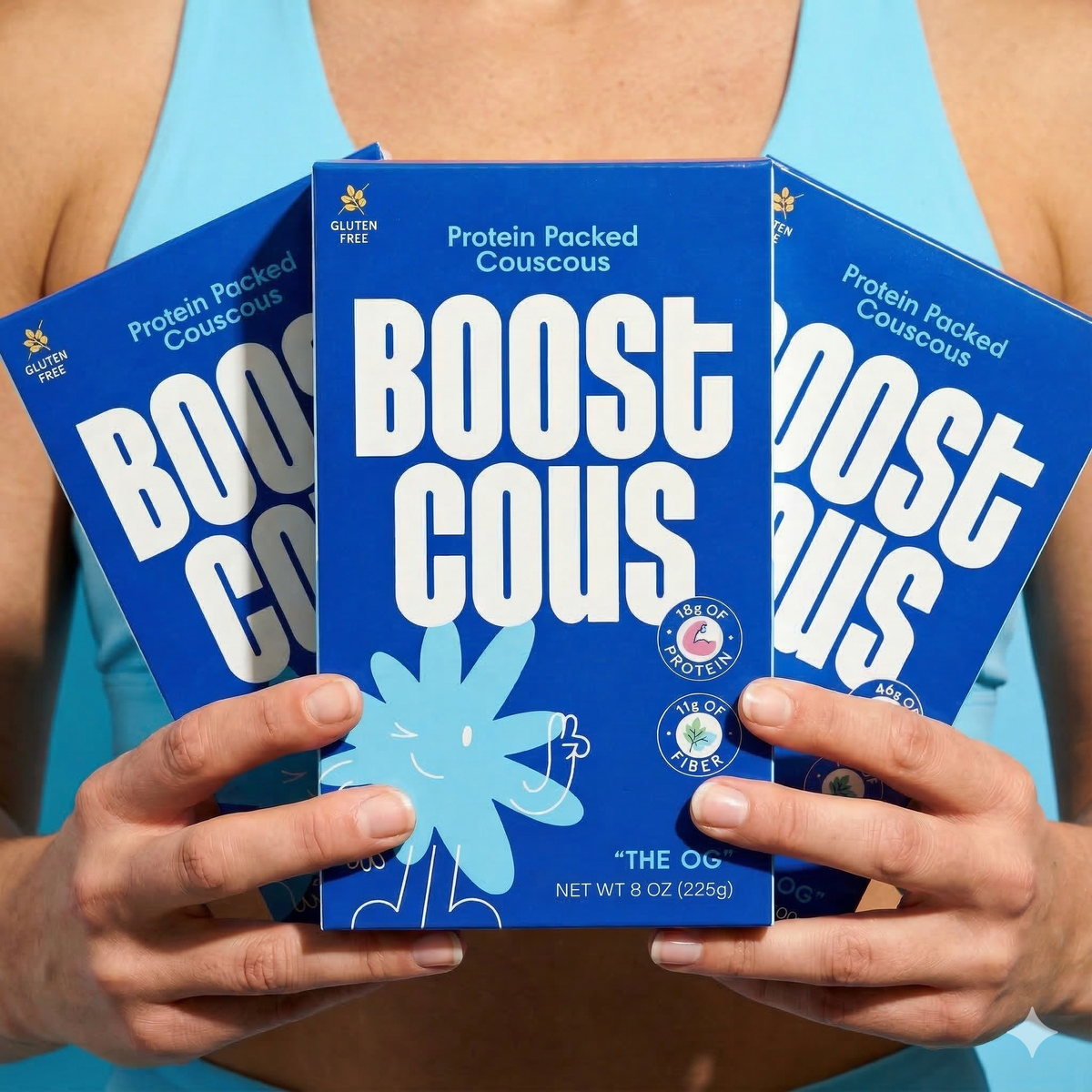 A person holds three boxes of "Boost Couscous," featuring bright blue packaging with playful graphics, showcasing the product's protein and gluten-free attributes against a light blue background.