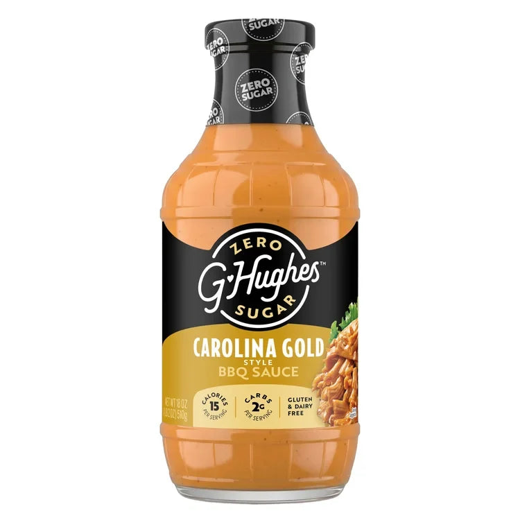 A clear bottle of G. Hughes Carolina Gold BBQ sauce features a golden-orange sauce, labeled as zero sugar, gluten-free, and low-calorie, with a serving suggestion of pulled pork on the side.