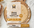 A package of Hero Flour Tortillas with six tortillas is displayed on a wooden cutting board, featuring nutritional information highlighting 16g of fiber and made with olive oil.