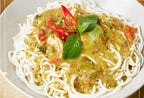 A bowl of noodles topped with a savory green sauce, diced vegetables, and red chili slices, garnished with fresh basil leaves, set on a light wooden surface.