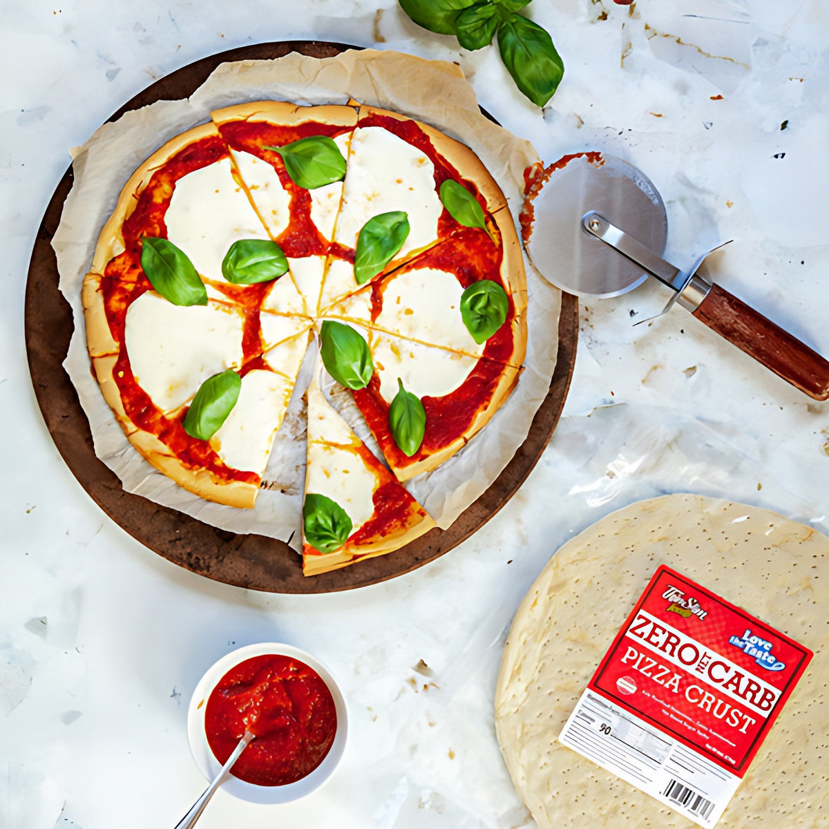 A pizza with tomato sauce, cheese, and basil beside a package of ThinSlim Foods Zero Net Carb Pizza Crust.