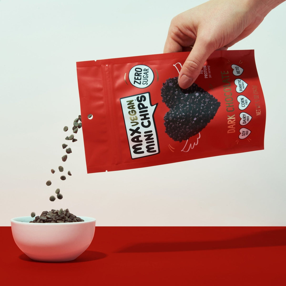 A hand pours dark chocolate mini chips from a red bag labeled "Max Vegan Mini Chips" into a white bowl against a simple background.