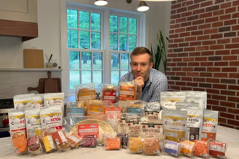 ThinSlim Foods founder Andrew Eckhardt behind a table of packaged breads, pasta, and sweets